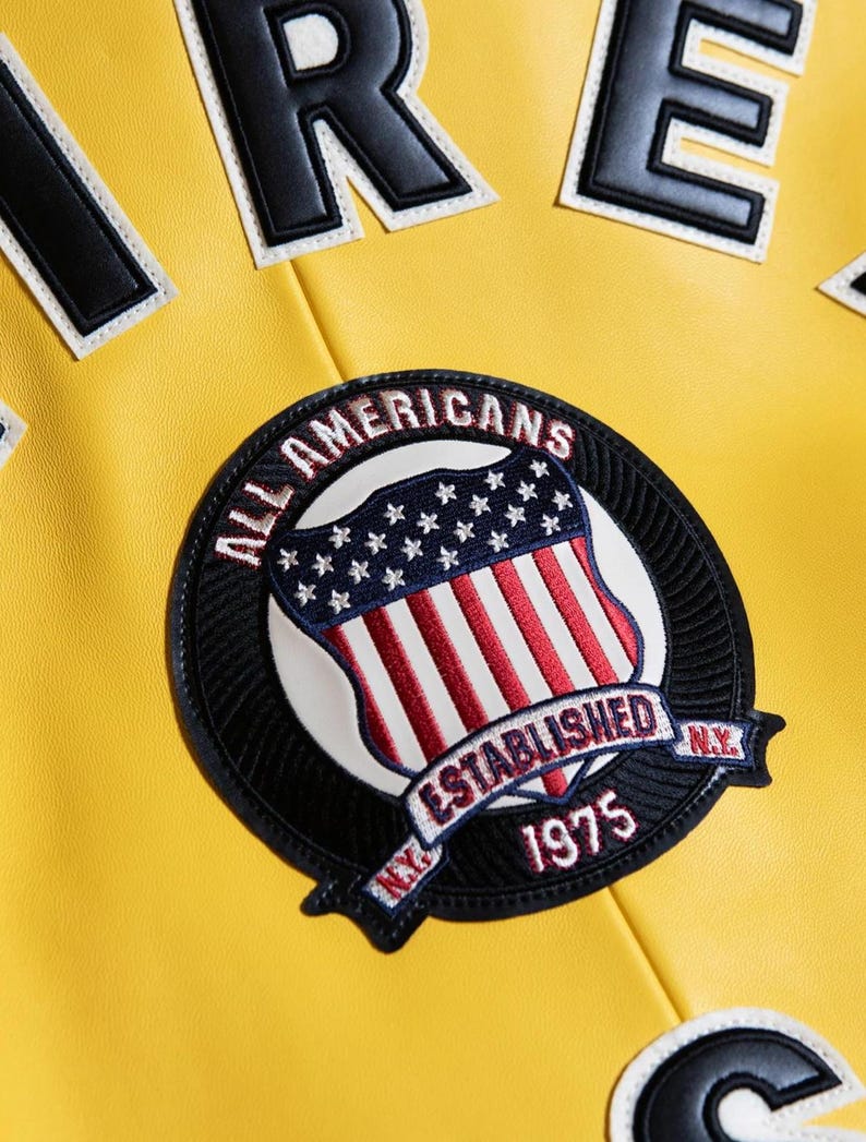 Pu&ograve; includere: Una giacca giallo brillante con una grande toppa circolare ricamata raffigurante uno scudo con strisce rosse e bianche e stelle blu. Il testo recita ALL AMERICANS ESTABLISHED 1975 N.Y. In alto sono visibili grandi lettere a blocchi bianche e nere.
