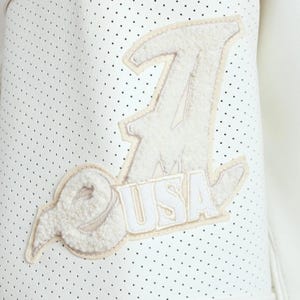 May include: A white perforated leather jacket sleeve featuring a large, textured, off-white chenille patch. The patch displays a stylized letter A with the text USA underneath, outlined in a light beige border against the white dotted material.