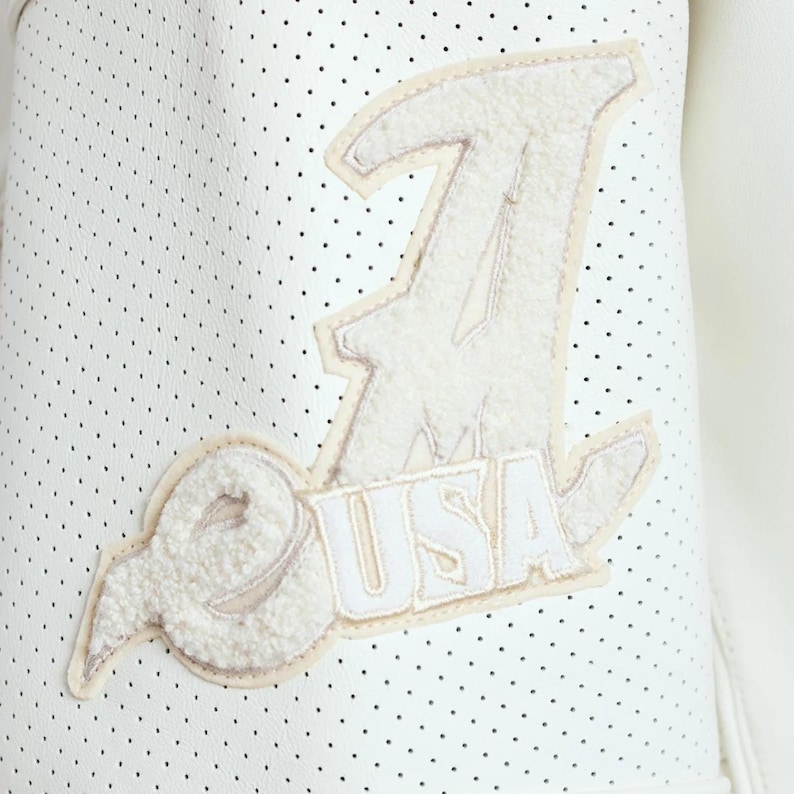 May include: A white perforated leather jacket sleeve featuring a large, textured, off-white chenille patch. The patch displays a stylized letter A with the text USA underneath, outlined in a light beige border against the white dotted material.