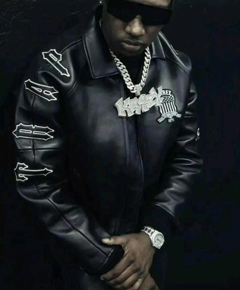 May include: A black leather jacket featuring the word TRAP embroidered in white on the right sleeve. The jacket includes a large silver-toned pendant necklace with the text KRAZY and a shield emblem with the text REX ESTABLISHED 1975.
