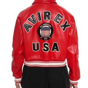 May include: A bright red jacket featuring the text AVIREX in black letters across the upper back and USA in black letters below a circular patch. The patch displays an American flag with the text ALL AMERICANS ESTABLISHED 1975. The jacket has white and red striped ribbed cuffs and a matching hem.