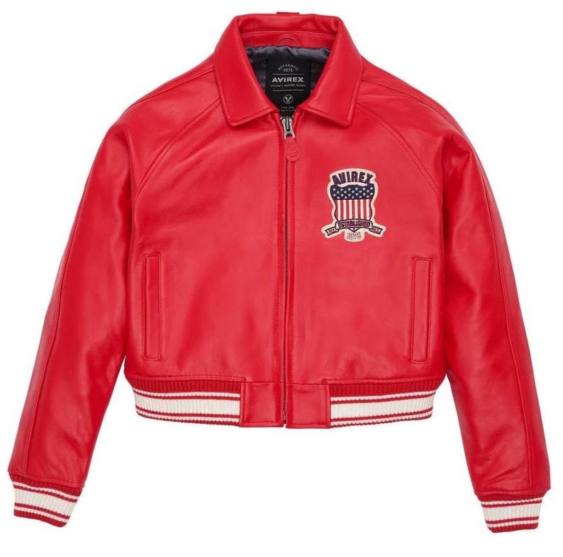 May include: A bright red leather bomber jacket featuring a classic collar, a full-length front zipper, and ribbed cuffs and hem with white stripes. A patch on the left chest displays the text AVIREX above a shield with stars and stripes.
