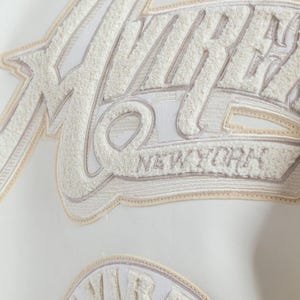 May include: A white jacket featuring large, textured cream-colored chenille lettering that spells out VIBES and NEW YORK. The embroidery is layered with beige and white stitching, creating a dimensional effect on the smooth white material surface.