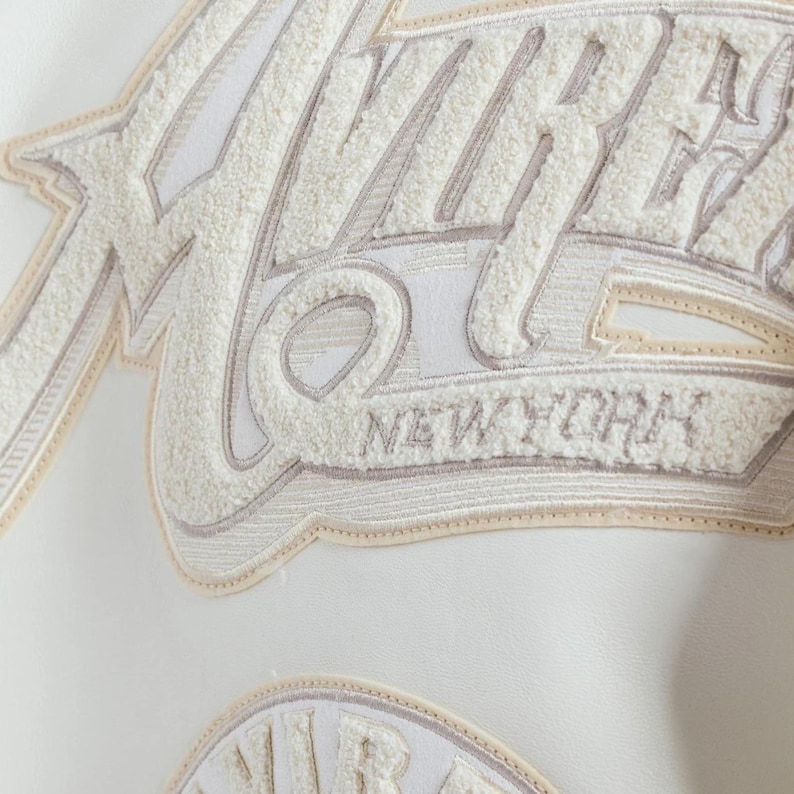 May include: A white jacket featuring large, textured cream-colored chenille lettering that spells out VIBES and NEW YORK. The embroidery is layered with beige and white stitching, creating a dimensional effect on the smooth white material surface.