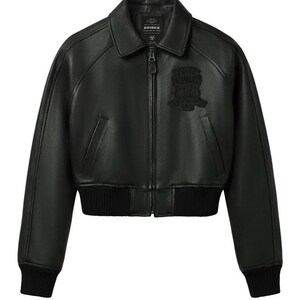 May include: A black leather bomber jacket featuring a classic collar, a full-length front zipper, and ribbed knit cuffs and hem. The chest displays a tonal embroidered patch with the text AVIREX. The jacket has two slanted side pockets.