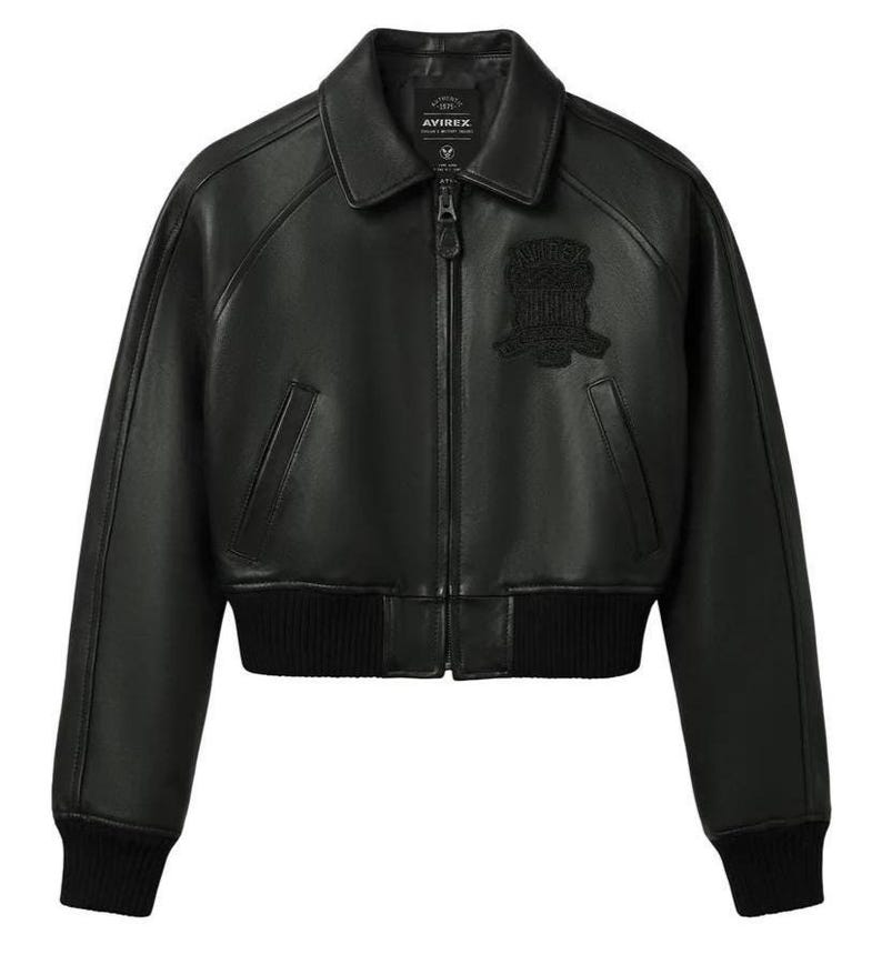 May include: A black leather bomber jacket featuring a classic collar, a full-length front zipper, and ribbed knit cuffs and hem. The chest displays a tonal embroidered patch with the text AVIREX. The jacket has two slanted side pockets.