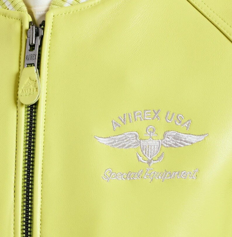 May include: A lime green leather jacket featuring a silver metal zipper with an Avirex branded pull tab. The chest displays white embroidered text reading AVIREX USA and Special Equipment above a winged shield and anchor emblem design.