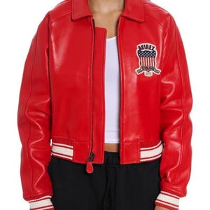 May include: A bright red leather bomber jacket featuring a classic collar, a front zipper closure, and white and red striped ribbed cuffs and hem. On the left chest, there is a patch with the text AVIREX and a graphic of the American flag.