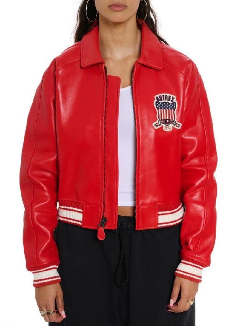 May include: A bright red leather bomber jacket featuring a classic collar, a front zipper closure, and white and red striped ribbed cuffs and hem. On the left chest, there is a patch with the text AVIREX and a graphic of the American flag.