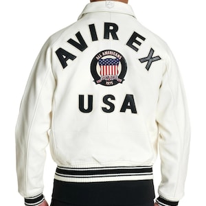 Puede incluir: Una chaqueta universitaria de cuero color blanco hueso con letras negras que dicen AVIREX y USA en la espalda. Un parche circular en el centro muestra el texto ALL AMERICANS ESTABLISHED 1975 con una bandera de EE. UU. La chaqueta tiene pu&ntilde;os y dobladillo acanalados con rayas blancas y negras.
