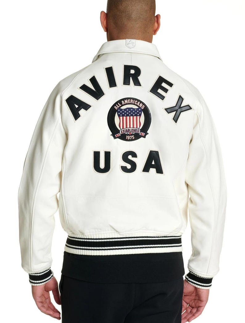 Puede incluir: Una chaqueta universitaria de cuero color blanco hueso con letras negras que dicen AVIREX y USA en la espalda. Un parche circular en el centro muestra el texto ALL AMERICANS ESTABLISHED 1975 con una bandera de EE. UU. La chaqueta tiene pu&ntilde;os y dobladillo acanalados con rayas blancas y negras.