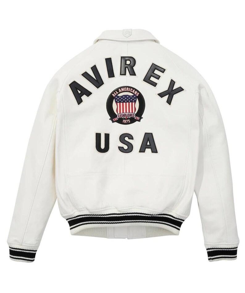 Puede incluir: Una chaqueta universitaria blanca con letras negras que dicen AVIREX y USA en la parte posterior. Un parche circular en el centro muestra una bandera estadounidense con el texto ALL AMERICANS ESTABLISHED 1975. La chaqueta tiene pu&ntilde;os acanalados a rayas.