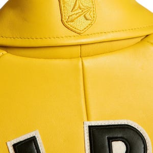 Pu&ograve; includere: Una giacca in pelle giallo brillante con colletto ripiegato e un piccolo emblema ricamato a forma di scudo giallo. Sotto il colletto, &egrave; visibile una parte di una grande lettera nera con bordo bianco sul retro del capo.