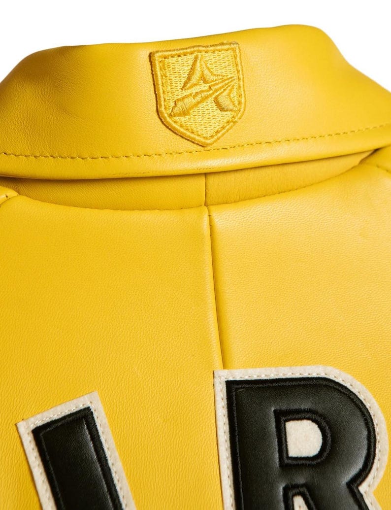 Pu&ograve; includere: Una giacca in pelle giallo brillante con colletto ripiegato e un piccolo emblema ricamato a forma di scudo giallo. Sotto il colletto, &egrave; visibile una parte di una grande lettera nera con bordo bianco sul retro del capo.