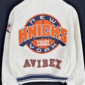Op de afbeelding: Een wit varsity-jack met een groot oranje en marineblauw New York Knicks basketbal-logo op de achterkant met de tekst KITH in het midden. Het woord AVIREX staat eronder in oranje en marineblauw. De mouwen en zoom hebben gestreepte geribbelde boorden.
