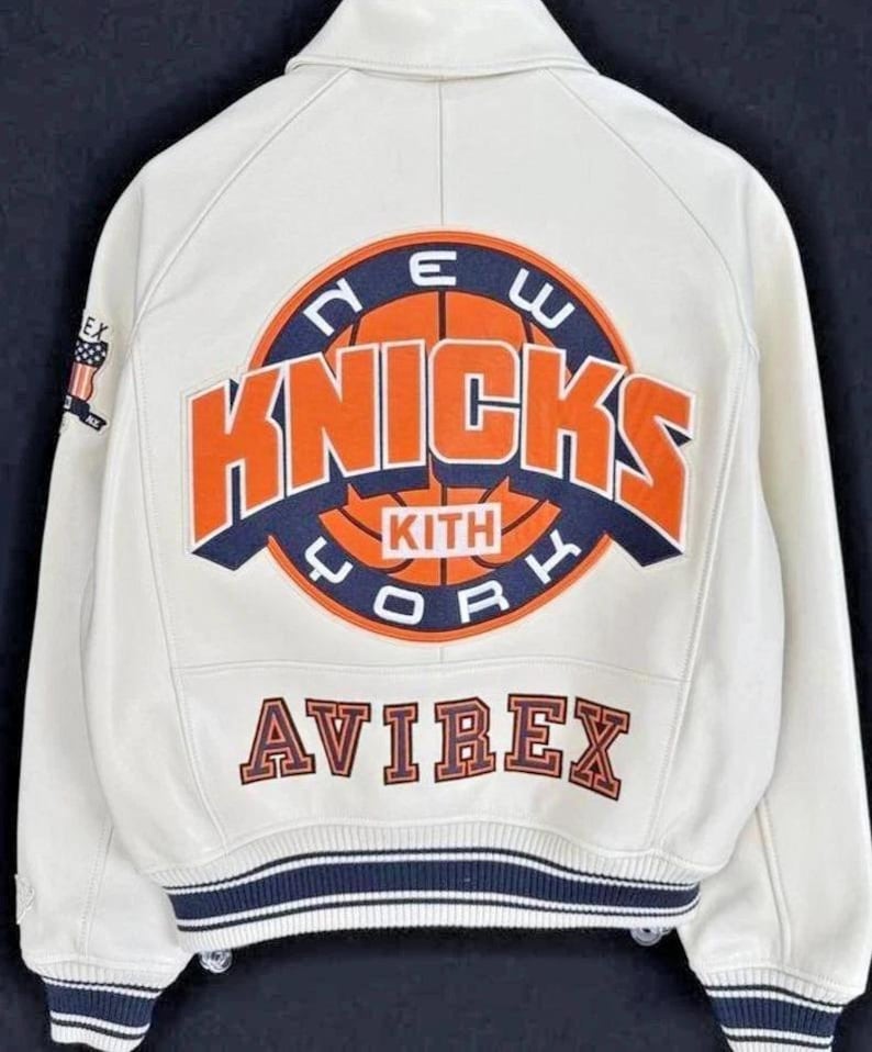 Op de afbeelding: Een wit varsity-jack met een groot oranje en marineblauw New York Knicks basketbal-logo op de achterkant met de tekst KITH in het midden. Het woord AVIREX staat eronder in oranje en marineblauw. De mouwen en zoom hebben gestreepte geribbelde boorden.