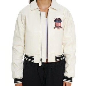 May include: An off-white leather bomber jacket featuring a front zipper closure, a pointed collar, and black and white striped ribbed cuffs and hem. A patch on the left chest displays the text AVIREX above a shield with red and white stripes.