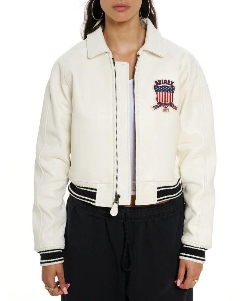 May include: An off-white leather bomber jacket featuring a front zipper closure, a pointed collar, and black and white striped ribbed cuffs and hem. A patch on the left chest displays the text AVIREX above a shield with red and white stripes.