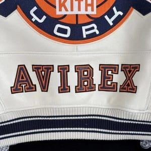 Op de afbeelding: Een wit leren varsity-jack met een grote geborduurde cirkelvormige patch met oranje en marineblauwe details, de tekst KITH en het woord YORK. Daaronder is het woord AVIREX geborduurd in marineblauw met oranje omlijningen.