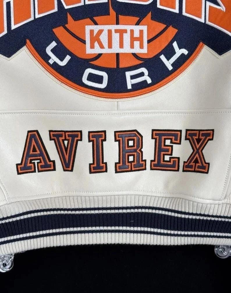 Op de afbeelding: Een wit leren varsity-jack met een grote geborduurde cirkelvormige patch met oranje en marineblauwe details, de tekst KITH en het woord YORK. Daaronder is het woord AVIREX geborduurd in marineblauw met oranje omlijningen.