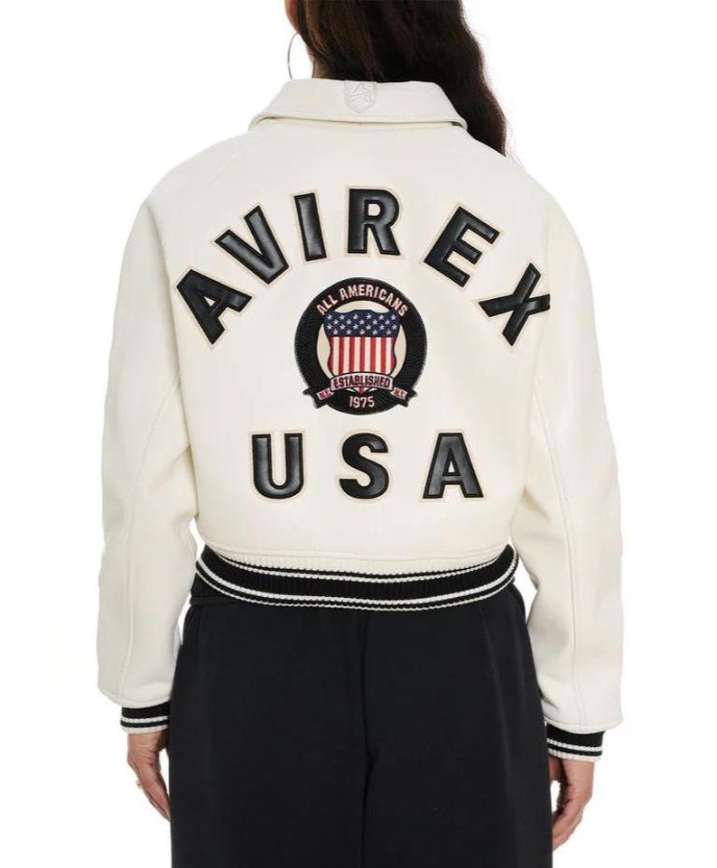 May include: An off-white varsity jacket featuring black lettering that reads AVIREX and USA on the back. A circular patch in the center displays a shield with the American flag, the text ALL AMERICANS, ESTABLISHED, and 1975. The jacket has striped trim.