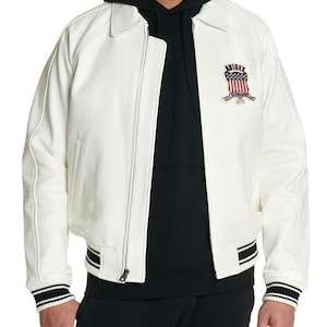 Puede incluir: Una chaqueta bomber de cuero blanco con cremallera frontal completa, cuello cl&aacute;sico y pu&ntilde;os y dobladillo acanalados con rayas negras y blancas. Un parche bordado en el pecho izquierdo muestra el texto AVIREX sobre un emblema de la bandera de EE. UU.