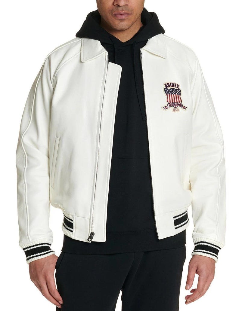 Puede incluir: Una chaqueta bomber de cuero blanco con cremallera frontal completa, cuello cl&aacute;sico y pu&ntilde;os y dobladillo acanalados con rayas negras y blancas. Un parche bordado en el pecho izquierdo muestra el texto AVIREX sobre un emblema de la bandera de EE. UU.