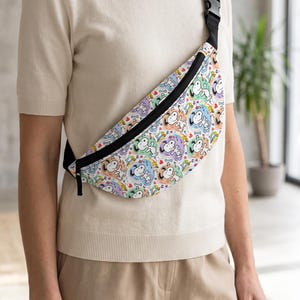 May include: A fanny pack featuring a repeating pattern of Snoopy characters in colorful circular frames against a white background. The bag has a black zipper closure and an adjustable black strap with a plastic buckle, worn across the torso.