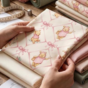 May include: A folded piece of cream-colored fabric featuring a repeating pattern of Winnie the Pooh wearing a pink shirt, interspersed with pink ribbon bows. The fabric is part of a larger stack of various solid and patterned textile rolls.