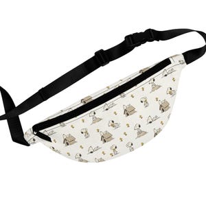 May include: A white fanny pack featuring an all-over print of the cartoon character Snoopy in various poses, including sitting on a doghouse and lying down, accented with small yellow flowers and a black adjustable strap with a plastic buckle.