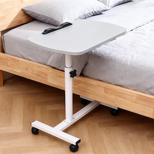 May include: A height-adjustable rolling bedside table with a light grey rectangular surface and a black plastic edge clip. The table features a white metal central support column with a black adjustment knob and a white base with four black wheels.