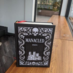 Puede incluir: Un libro de tapa dura negro con el título "MANACLED" y el nombre del autor "SENLINYU" en blanco. El diseño de la portada incluye una calavera, serpientes y una ilustración de un edificio gótico. Una cinta roja de marcador es visible.