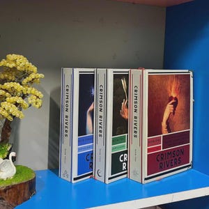Puede incluir: Cuatro libros de tapa dura titulados "Crimson Rivers" se exhiben en una estantería azul. Los lomos de los libros son blancos con el título en negro. Cada portada de libro presenta una ilustración artística diferente. Los libros forman parte de una serie.