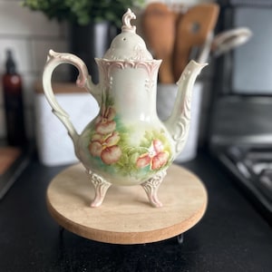 May include: A porcelain teapot featuring a cream-colored base with hand-painted pink and orange pansy floral motifs. The vessel includes an ornate handle, a curved spout, a removable lid with a finial, and three decorative feet with pink accents.