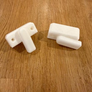Peut inclure: Deux objets blancs et rectangulaires imprimés en 3D. L'un a une base rectangulaire avec deux trous et une pièce rectangulaire plus petite perpendiculaire. L'autre a deux pièces rectangulaires, une plus grande et une plus petite.