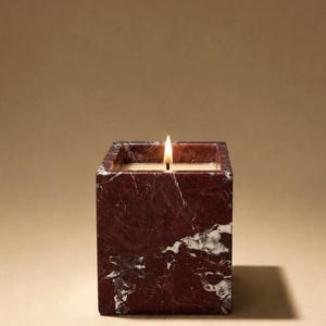 May include: A square candle holder made of deep red marble with white veining, containing a lit candle with a visible flame. The vessel has a smooth, polished finish and a geometric shape, showcasing the natural stone patterns throughout the exterior.