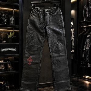 Puede incluir: Pantalones de cuero negro con cierre de botones y múltiples motivos de cruces en relieve en las piernas. Una cruz prominente en la parte inferior de la pierna izquierda es de color rojo, mientras que las demás son de color negro tonal.