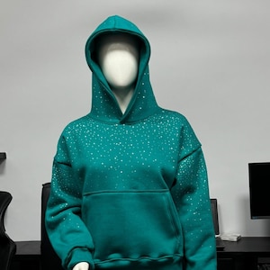 May include: A teal hooded sweatshirt featuring a scattered pattern of small, reflective silver dots across the front, sleeves, and hood. The garment includes a large front kangaroo pocket, long sleeves with ribbed cuffs, and a matching ribbed hem.