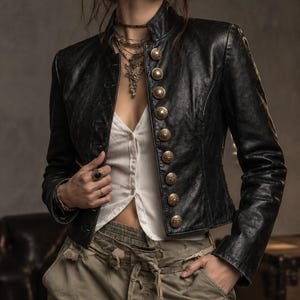 Puede incluir: Una chaqueta de cuero negro con cuello alto y una fila de botones metálicos grandes, dorados y ornamentados en la parte delantera. La chaqueta se lleva sobre una camisa blanca abotonada y pantalones verde oliva con cordón en la cintura.