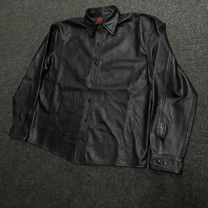 Puede incluir: Una camisa negra de manga larga con botones, hecha de un material liso similar al cuero. La camisa presenta un cuello en punta, una tapeta frontal con botones negros a juego y pu&ntilde;os abotonados. Un peque&ntilde;o logotipo rojo es visible en la etiqueta.