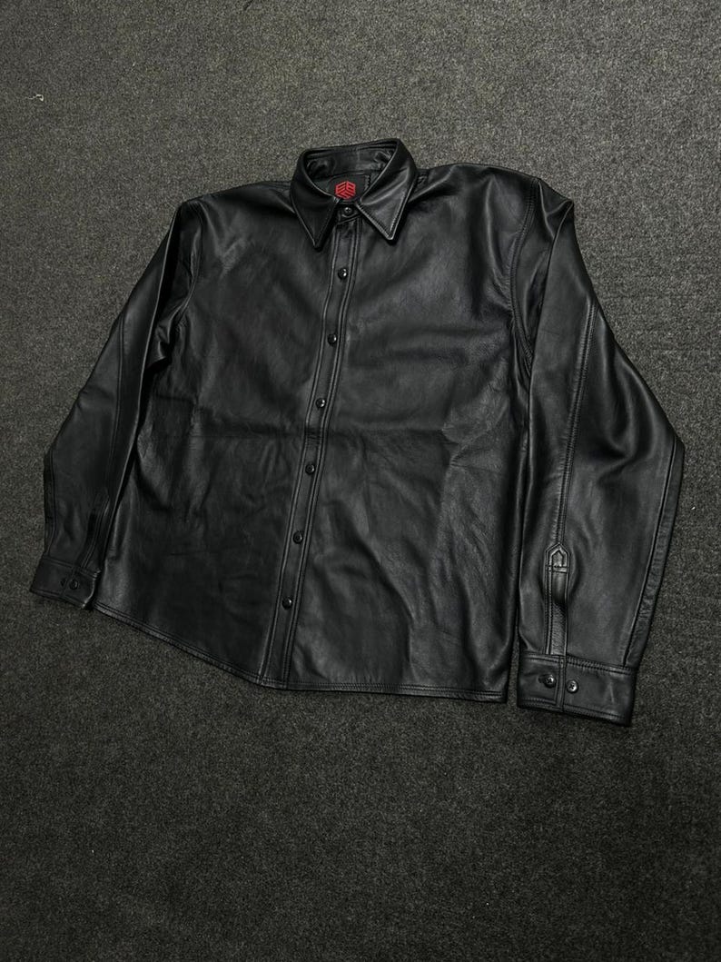 Puede incluir: Una camisa negra de manga larga con botones, hecha de un material liso similar al cuero. La camisa presenta un cuello en punta, una tapeta frontal con botones negros a juego y pu&ntilde;os abotonados. Un peque&ntilde;o logotipo rojo es visible en la etiqueta.