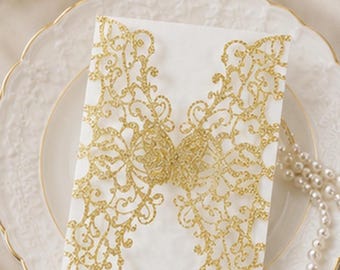 May include: A white invitation card with intricate gold glitter cut-out butterfly and floral designs. The card is displayed on a white plate with a gold rim, alongside a pearl necklace. Ideal for weddings or special events.