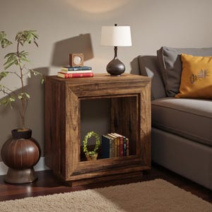 May include: A square, wooden side table with a dark brown finish. A lamp with a white shade sits on top, along with books and a small clock. The table has an open shelf with books and a small plant. A decorative accent for a living room.