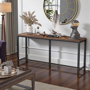May include: A long, rectangular console table with a dark brown, wood-look top and black metal frame. The table is decorated with a vase of dried flowers, decorative objects, and a sculpture. The table is 60 inches long.