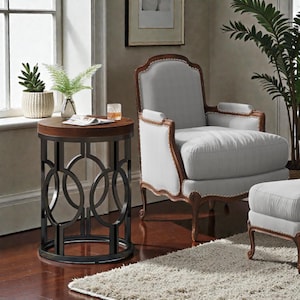 May include: A round side table with a dark wood top and a black metal base featuring a geometric design. The table is next to a white upholstered armchair with a wooden frame. A small rug and a potted plant are also visible.