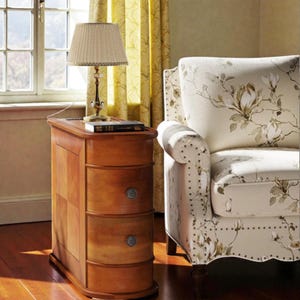May include: A wooden nightstand with three drawers and a lamp. The nightstand is a warm brown color with a curved front and silver drawer pulls. A beige lamp with a pleated shade sits on top, next to a book. A floral armchair is to the right.