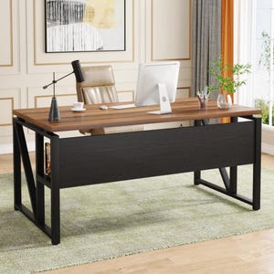 May include: A rectangular desk with a dark wood grain top and a black metal frame. The desk features a black modesty panel, a black desk lamp, a white computer monitor, a beige office chair, and a small glass vase with green plant stems.