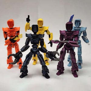 Könnte beinhalten: Eine Gruppe von fünf bunten Roboter-Actionfiguren. Die Roboter sind in den Farbtönen Orange, Gelb, Schwarz, Lila und Blau gehalten. Jede Figur hat ein einzigartiges Design mit beweglichen Gliedmaßen und verschiedenen waffenartigen Aufsätzen. Die Figuren stehen auf einer weißen Oberfläche.