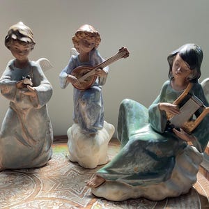 May include: Three porcelain angel figurines with glossy finishes. One angel stands holding a small bird, another stands on a cloud playing a lute, and the third sits on a cloud playing a harp. The figures feature muted blue, green, and white tones.