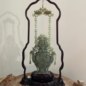 May include: A pale green carved jade vessel suspended by two carved jade chains from a dark brown wooden frame. The vessel features intricate relief carvings of birds and mythical creatures, resting on a matching dark brown carved wooden base.